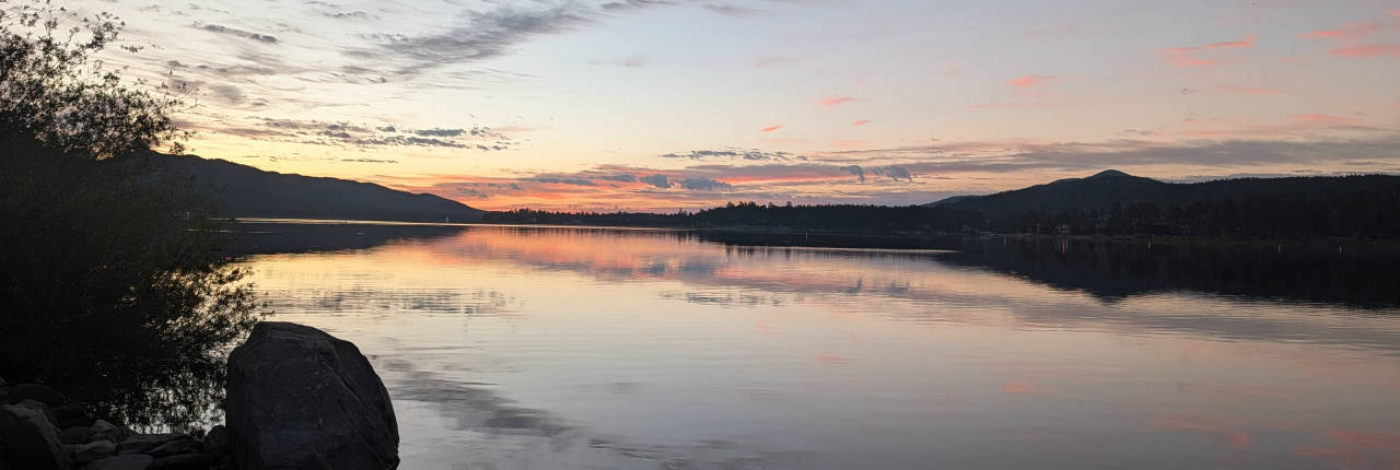Big Bear Lake at sunset, pink cloud reflected in still water with a boulder in the foreground