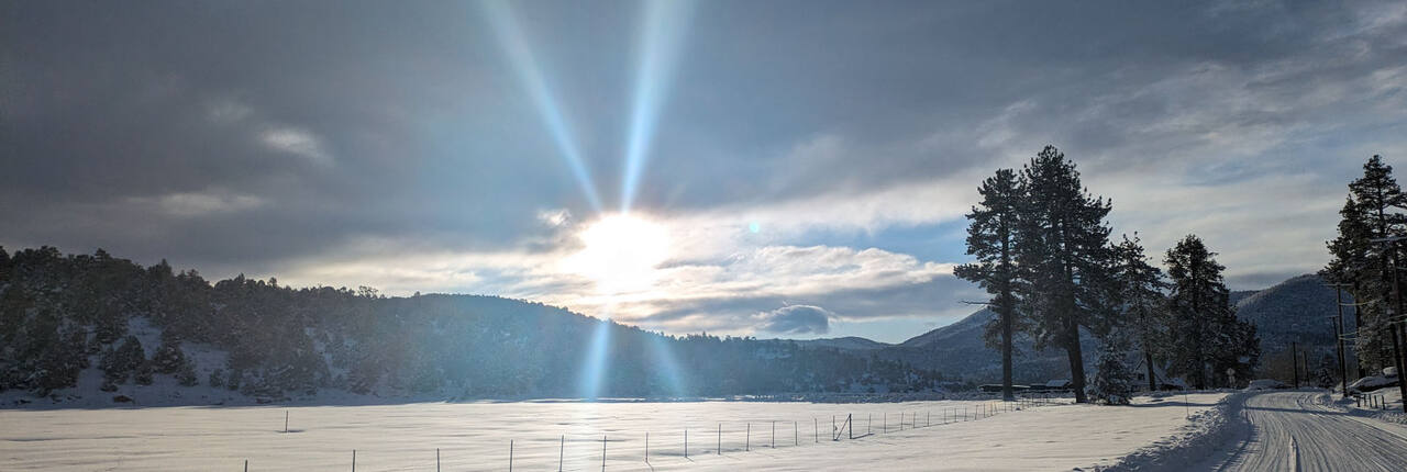 Sun breaking through cloud over a snow-covered Big Bear Lake, with a plowed road running along the shoreline